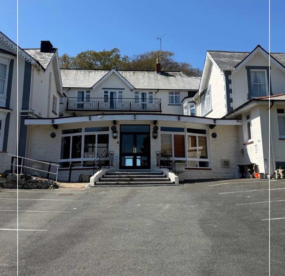 Front view of The Queens Road Hotel with main entrance, curved canopy, and white facade under a clear blue sky